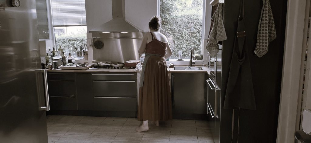 Noor staat met haar rug naar de camera in een keuken te werken, ze heeft een lange oranje rok aan en blote voeten en haar haar zit in een knot