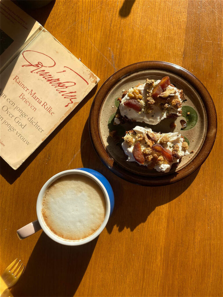 Vegan bananenbrood getopt met yoghurt en gedroogd fruit en noten, op een bordje, met daarnaast een cappuchino en een boek: 'brieven'- van Rainer Maria Rilke