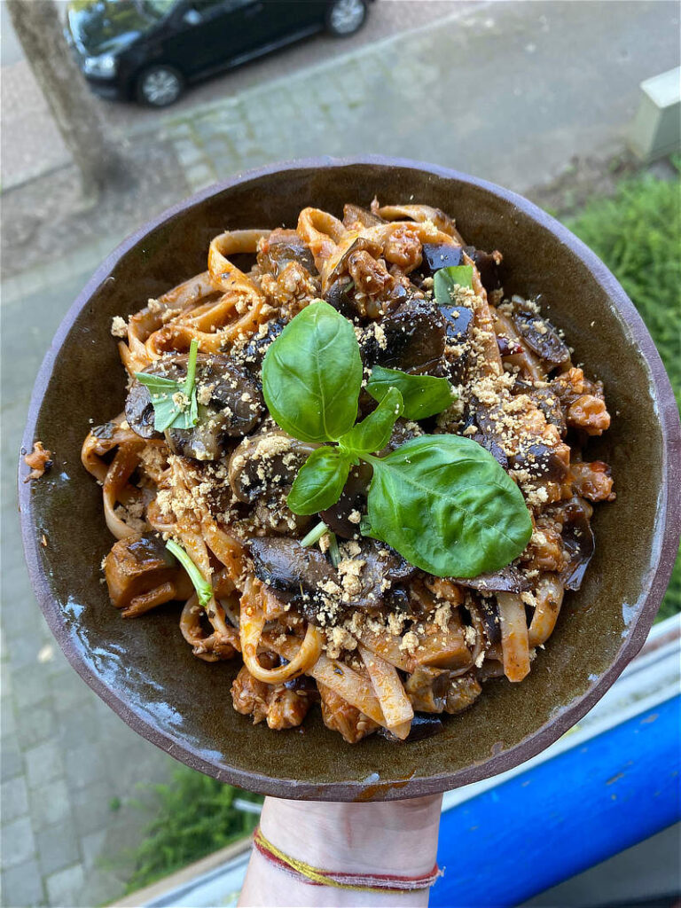 Tagliatelle met paddenstoelen en walnoten, met een groot blad basilicum er op, in een bruin schaaltje wat vastgehouden wordt in 1 hand