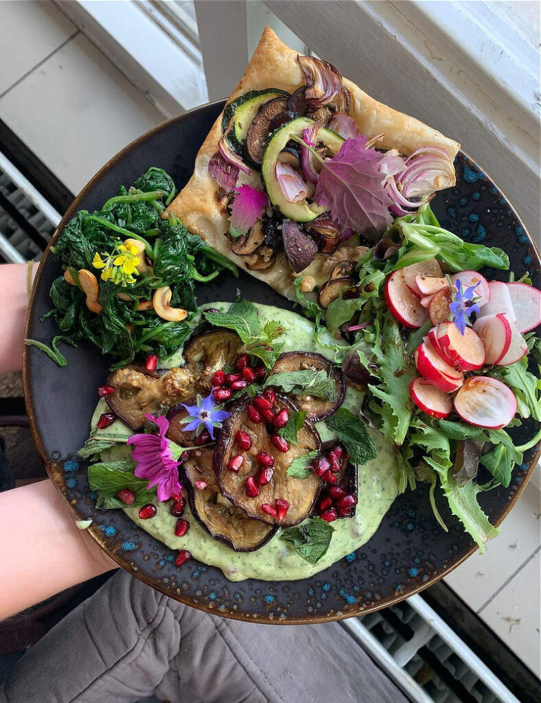 veganistische maaltijd op een bord, bestaande uit salade, een stuk plaattaart, yoghurtsaus met aubergines erop en een beetje spinazie, versierd met kleurrijke eetbare bloemetjes