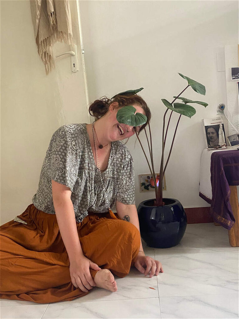 een vrolijke foto van Noor de Vries, vrouw 24, zittend op de grond met een kamerplant waar haar gezicht speels achter verstopt is